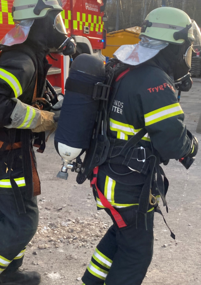 2 brandkadetter i fuld uniform. De er parat til røgdykning med hjelme, masker og iltmasker på ryggen.